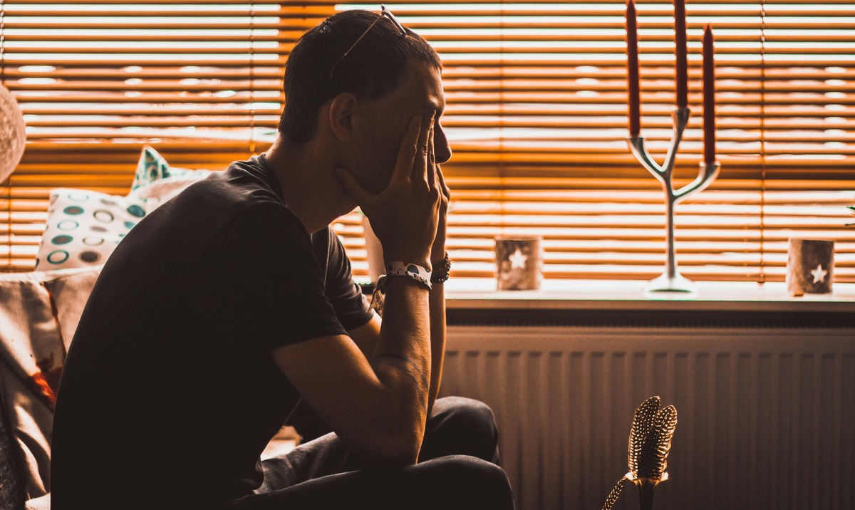 A man sits alone beside a window with his hand pressed over his face, showing the bodily tension that anxiety can produce alongside worry: tight muscles, shallow breathing and disturbed sleep.