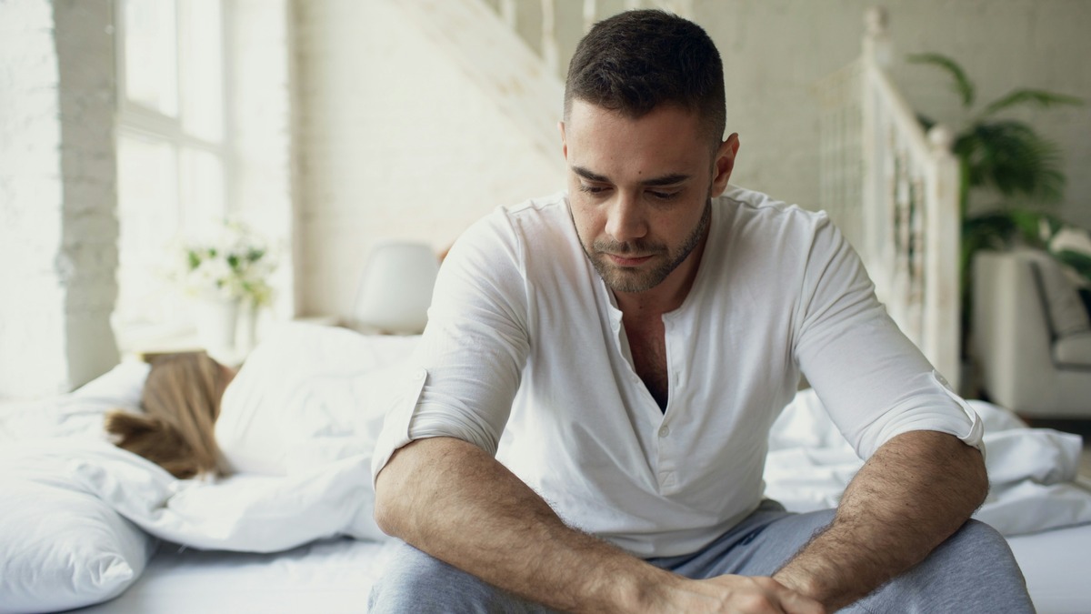 A man sits on the edge of his bed with his head lowered while his partner sleeps behind him, reflecting the low mood, stress and sleep difficulties men commonly seek mental health care for at Helyx Health.