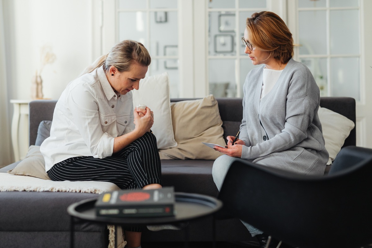 A woman sits forward with her hands clasped while her therapist listens and makes notes beside her, a session of the anxiety therapy Helyx Health coordinates.