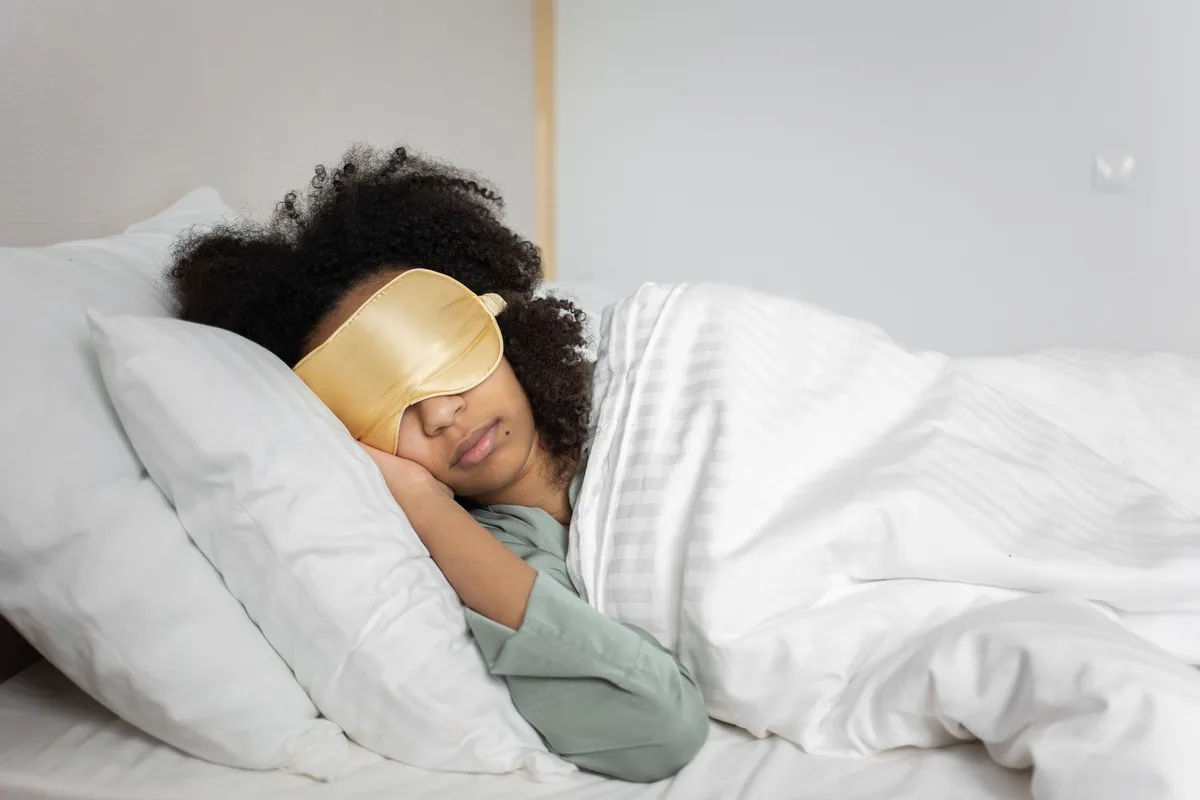 A woman sleeps on her side in a bright bedroom wearing a silk eye mask, the rest a sleep study is arranged to measure and explain.